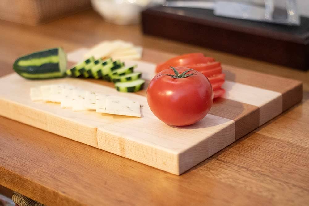 Maple and cherry wooden cutting board with sliced tomatoes, cucumber, and cheeses