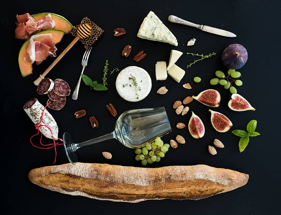 Wine and snack set. Baguette, glass of white, figs, grapes, nuts, cheese variety, meat appetizers and herbs on black background, top view